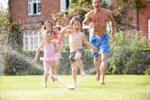 Family running and jumping through garden sprinkler